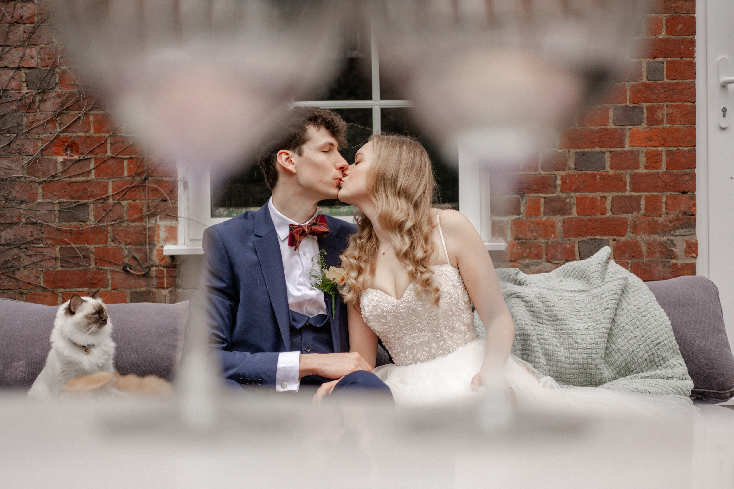 A newly married couple shares a kiss while their cat watches, with two blurred champagne glasses in the foreground.